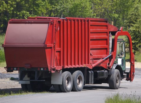 A large red commercial waste collection truck parked on a paved surface, positioned to the right side of the image. The truck's cargo container is prominently displayed, featuring a smooth, painted metal surface with vertical reinforced panels and a hydraulic lifting mechanism attached to the rear for emptying bins. The cabin is located at the front, with a white side mirror visible on the driver's side. The background includes a natural setting with green trees and foliage, indicating an outdoor environment. The scene is well-lit with natural daylight, emphasizing the vehicle's bold red colour and the textured surface of the cargo area, suggesting it is prepared for rubbish removal tasks typical of commercial waste services in Southfields or nearby areas.