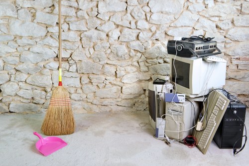 The image depicts a collection of old electronic waste and household items placed on a concrete floor against a textured beige stone wall. Visible items include an outdated computer monitor with a CRT screen, a beige computer tower, a small printer, and a wired keyboard. Nearby, there is an electric fan resting on the ground, along with tangled cords and cables draped over the electronic devices. To the left of the electronic clutter, a pink plastic dustpan and a small yellow-handled broom with a natural bristle sweeping surface are positioned against the wall. The scene suggests an indoor or enclosed outdoor area suitable for waste collection or disposal, with the arrangement indicating a recent clearing of unwanted items in preparation for rubbish removal services, such as those offered by Commercial Waste Southfields in the Southfields area, close to London SW18 postcode.
