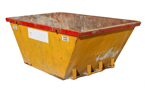 A large, yellow industrial skip with a red reinforced top edge is positioned outdoors on a concrete surface, likely on a driveway or road, as part of waste removal activities. The skip's metal walls show signs of use, with visible patches of dirt, rust, and chipped paint, indicating frequent servicing. The interior is also rusty and appears to contain residual debris. The skip is rectangular with a tapered design, wider at the top and narrower at the base, stabilized by four yellow support brackets at the bottom corners. In the background, there are no other objects or scenery visible, suggesting the skip is situated in an open space typical of commercial waste collection in Southfields or wider London areas. This scene aligns with waste management services by companies like Commercial Waste Southfields, who handle large-scale rubbish removal for local businesses and construction sites, emphasizing the importance of proper disposal methods for bulky waste materials.