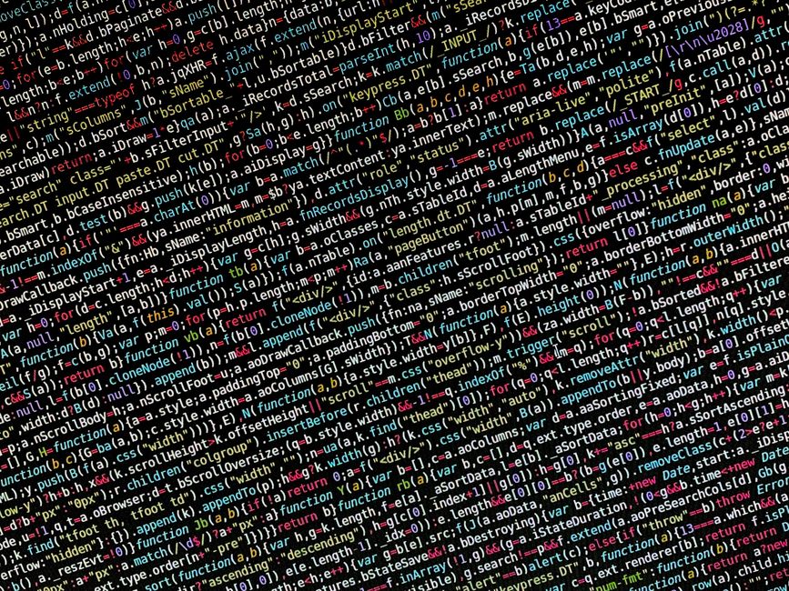 A person working at a desk with a laptop displaying colorful lines of code on the screen, indicative of programming or software development. The individual is wearing a light blue shirt and is holding